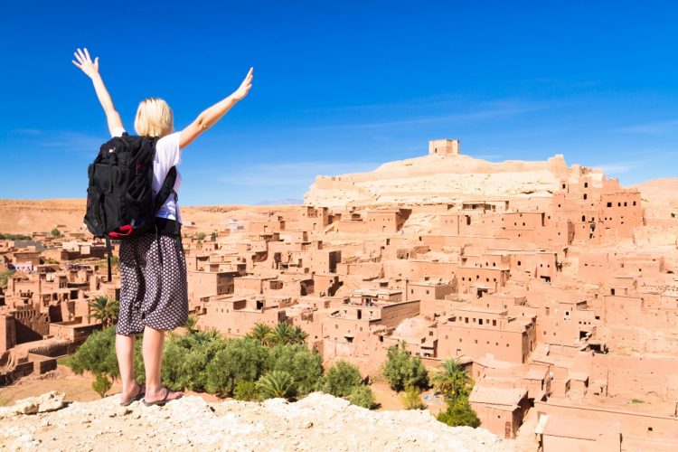 Adventurous-woman-arms-reised-in-front-of-Ait-Benhaddou-fortified-city-kasbah-or-ksar-along-the-former-caravan-route-between-Sahara-and-Marrakesh-in-present-day-Morocco